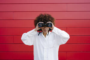 Afro-Frau schaut durch ein Fernglas vor einer roten Wand - GIOF15533