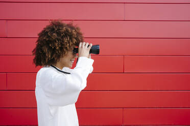 Smiling young woman looking through binoculars by red wall - GIOF15534