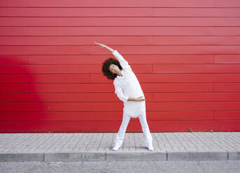 Young woman stretching hands on footpath in front of red wall - GIOF15538