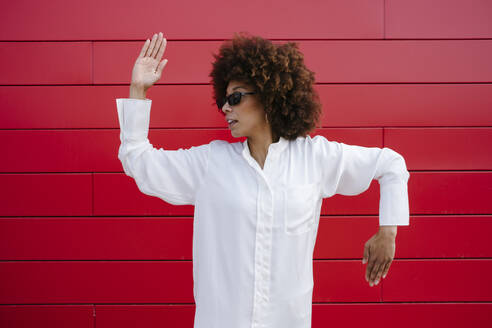 Afro junge Frau mit Sonnenbrille tanzt vor einer roten Wand - GIOF15540