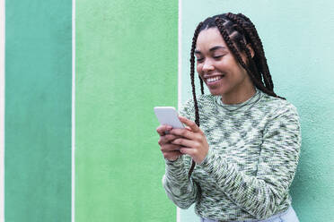 Lächelnde junge Frau, die ein Smartphone benutzt und vor einer Wand steht - PNAF03900
