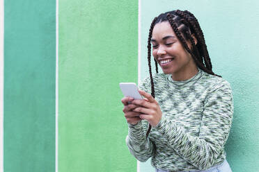 Smiling young woman using smart phone standing in front of wall - PNAF03900