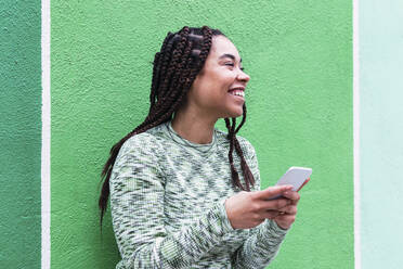 Happy young woman with smart phone standing in front of green wall - PNAF03902