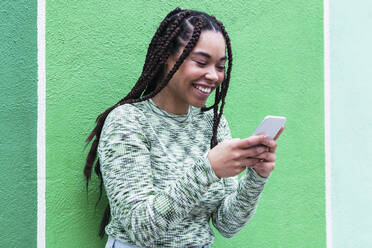 Happy woman using smart phone standing in front of green wall - PNAF03903