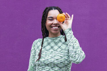 Happy woman covering eye with orange in front of purple wall - PNAF03906