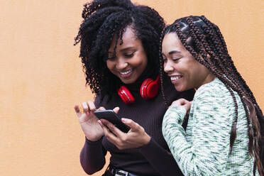 Happy woman sharing smart phone with friend standing in front of wall - PNAF03911