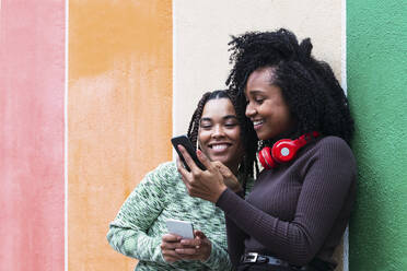 Happy woman with friend using smart phone in front of wall - PNAF03916