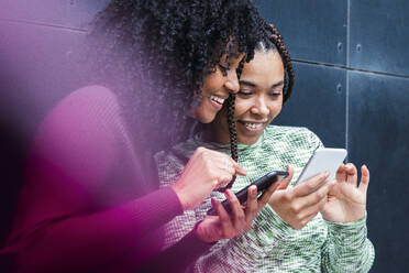 Smiling female friends using smart phones in front of wall - PNAF03935