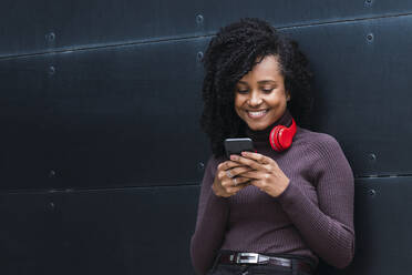 Lächelnde Frau mit drahtlosen Kopfhörern und Smartphone vor einer Wand - PNAF03937