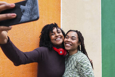 Fröhliche Frau mit Freund, die ein Selfie mit ihrem Smartphone vor einer Wand macht - PNAF03950