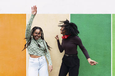 Happy female friends enjoying dance in front of multi colored wall - PNAF03957