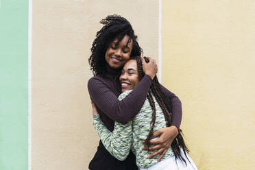 Happy female friends hugging each other standing in front of wall - PNAF03961