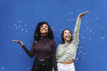 Happy friends throwing confetti standing in front of blue wall - PNAF03972