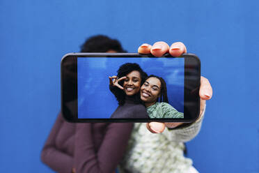 Happy friends taking selfie through smart phone in front of blue wall - PNAF03985