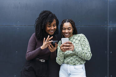 Happy young woman with friend using mobile phones in front of wall - PNAF03987