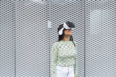 Smiling woman wearing virtual reality simulator standing in front of metal wall - PNAF03992