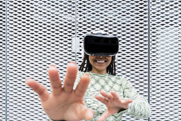 Lächelnde junge Frau in einem Virtual-Reality-Simulator gestikuliert vor einer Metallwand - PNAF03993