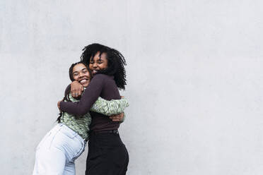 Cheerful female friends hugging each other in front of wall - PNAF04011