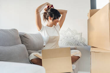 Happy woman tying hair sitting with cardboard box on sofa in living room at home - EGHF00413