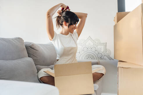Happy woman tying hair sitting with cardboard box on sofa in living room at home - EGHF00413