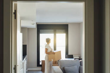 Woman with cardboard box standing in living room at new home - EGHF00431
