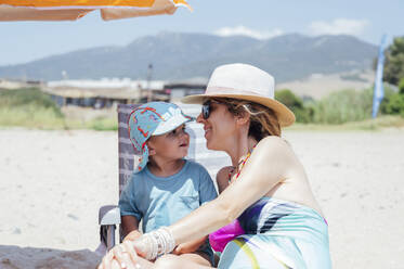 Smiling woman wearing hat sitting with son at beach on sunny day - PGF01115
