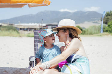 Happy mother wearing hat sitting with son at beach on sunny day - PGF01116