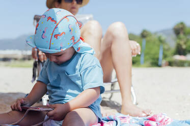 Junge benutzt Mobiltelefon vor der Mutter, die sich an einem sonnigen Tag am Strand entspannt - PGF01117