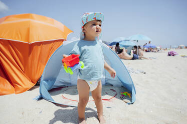 Boy holding toy car standing in front of tent at beach on sunny day - PGF01121