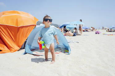 Niedlicher Junge mit Sonnenbrille, der mit einem Spielzeugauto vor einem Zelt am Strand steht - PGF01122