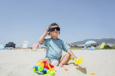 Boy adjusting sunglasses sitting with toys on sand at beach - PGF01123