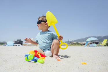 Boy wearing sunglasses holding shovel at beach on sunny day - PGF01124