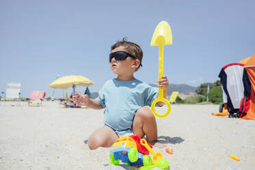 Süßer Junge mit Sonnenbrille spielt mit Spielzeug am Strand an einem sonnigen Tag - PGF01125