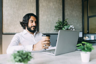 Businessman with coffee up using laptop at office - MEUF06065