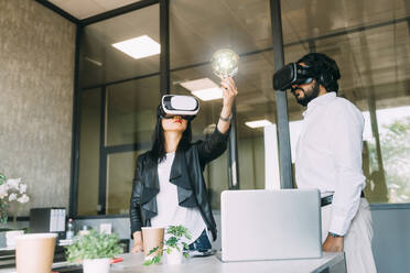 Businesswoman wearing VR glasses and examining light bulb by colleague - MEUF06084