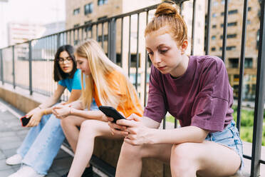 Mädchen mit blondem Haar, das ein Smartphone benutzt und mit Freunden vor einem Geländer sitzt - MEUF06172