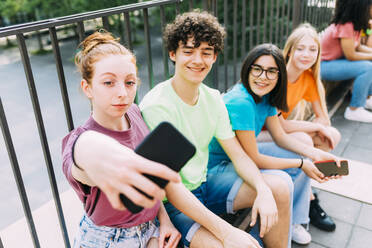 Lächelndes Teenager-Mädchen, das ein Selfie mit Freunden macht, die am Geländer sitzen - MEUF06175