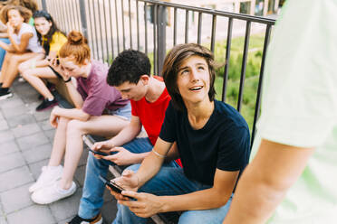Junge mit Handy sitzt bei Freunden vor dem Geländer - MEUF06186