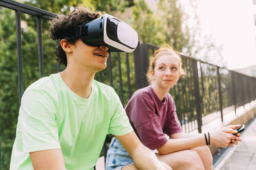 Lächelnder Junge, der einen Virtual-Reality-Simulator trägt und mit einem Freund vor einem Geländer sitzt - MEUF06215