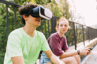 Smiling boy wearing virtual reality simulator sitting by friend in front of railing - MEUF06215