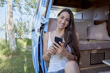 Lächelnde Frau mit Smartphone hört Musik über In-Ear-Kopfhörer im Wohnmobil - FMOF01475