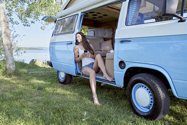 Woman with mobile phone day dreaming in motor home by lake - FMOF01476