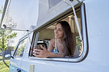 Smiling woman with coffee cup looking out of camper van window - FMOF01478