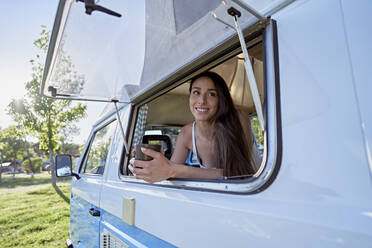 Woman with coffee cup looking through window of motor home - FMOF01479