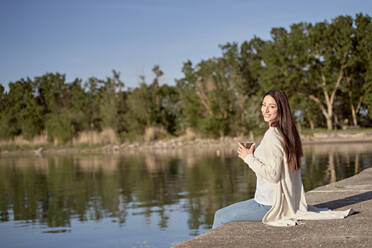 Smiling woman with coffee cup sitting on jetty by lake - FMOF01488