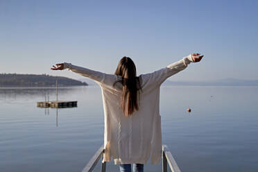 Woman standing with arms outstretched at lake on sunny day - FMOF01489