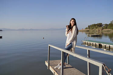 Junge Frau mit Kaffeetasse auf einem Steg über dem See stehend - FMOF01492