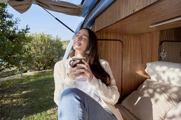 Smiling woman with coffee cup day dreaming in camper van - FMOF01496