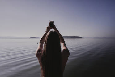 Junge Frau beim Fotografieren mit dem Handy am See - FMOF01501