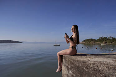 Lächelnde Frau mit Smartphone auf einem Steg am See - FMOF01502